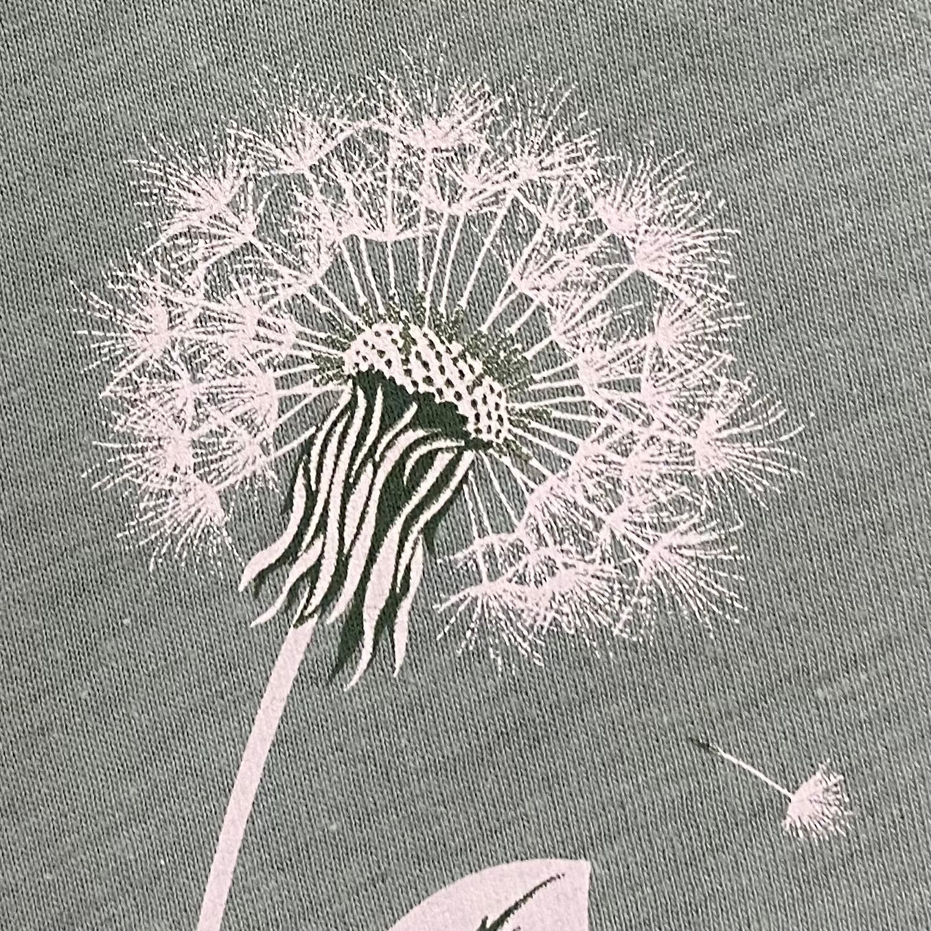 Detailed, silhouette of a dandelion seed blowing in the wind on a super soft, heather dusty green nature inspired  graphic tee .