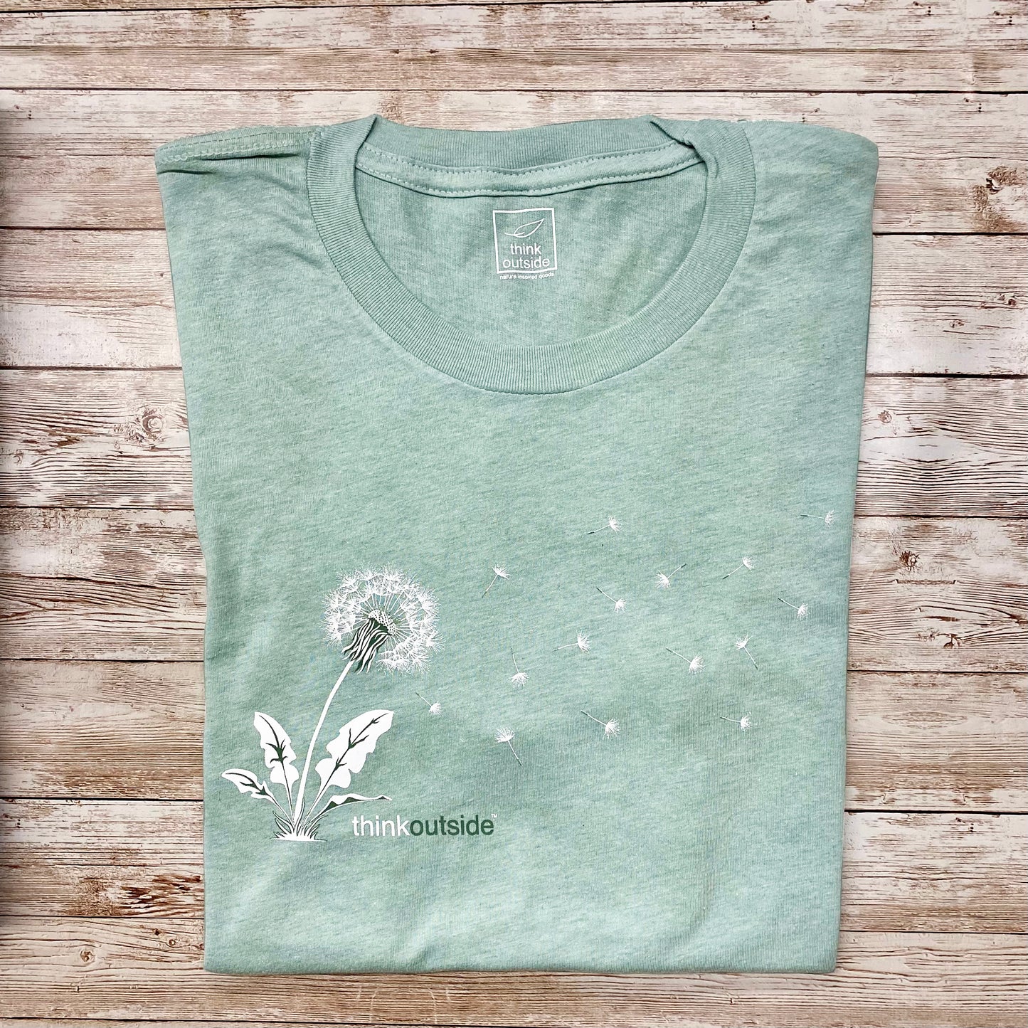 Hand drawn silhouette of a dandelion seed blowing in the wind on a super soft, heather dusty green nature inspired graphic tee .