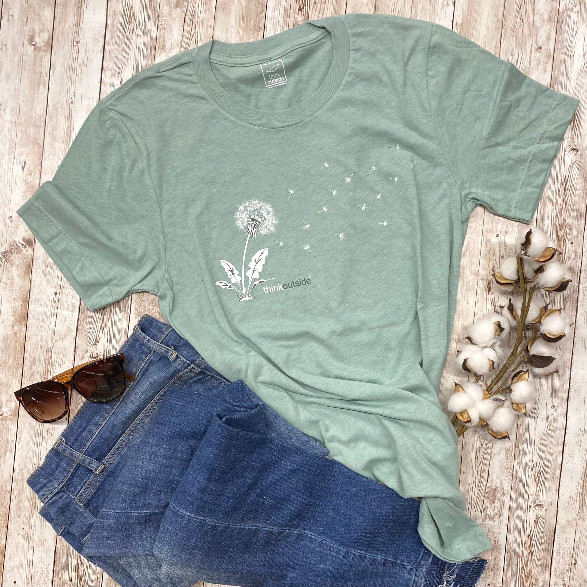 A super soft, heather dusty green  gardening graphic tee with a silhouette of a dandelion seed blowing in the wind for the outdoor lover. Features the phrase Think Outside.
