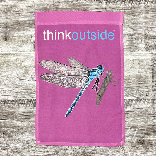 Blue dragonfly with lacy wings clutching its shell on a bright purplish pink garden flag with the phrase Think Outside.