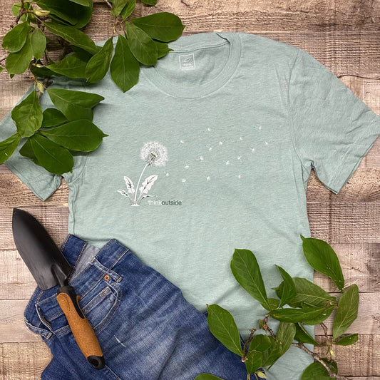 Silhouette of a dandelion seed blowing in the wind on a super soft, heather dusty green outdoor gardening graphic tee. Features the phrase Think Outside. 