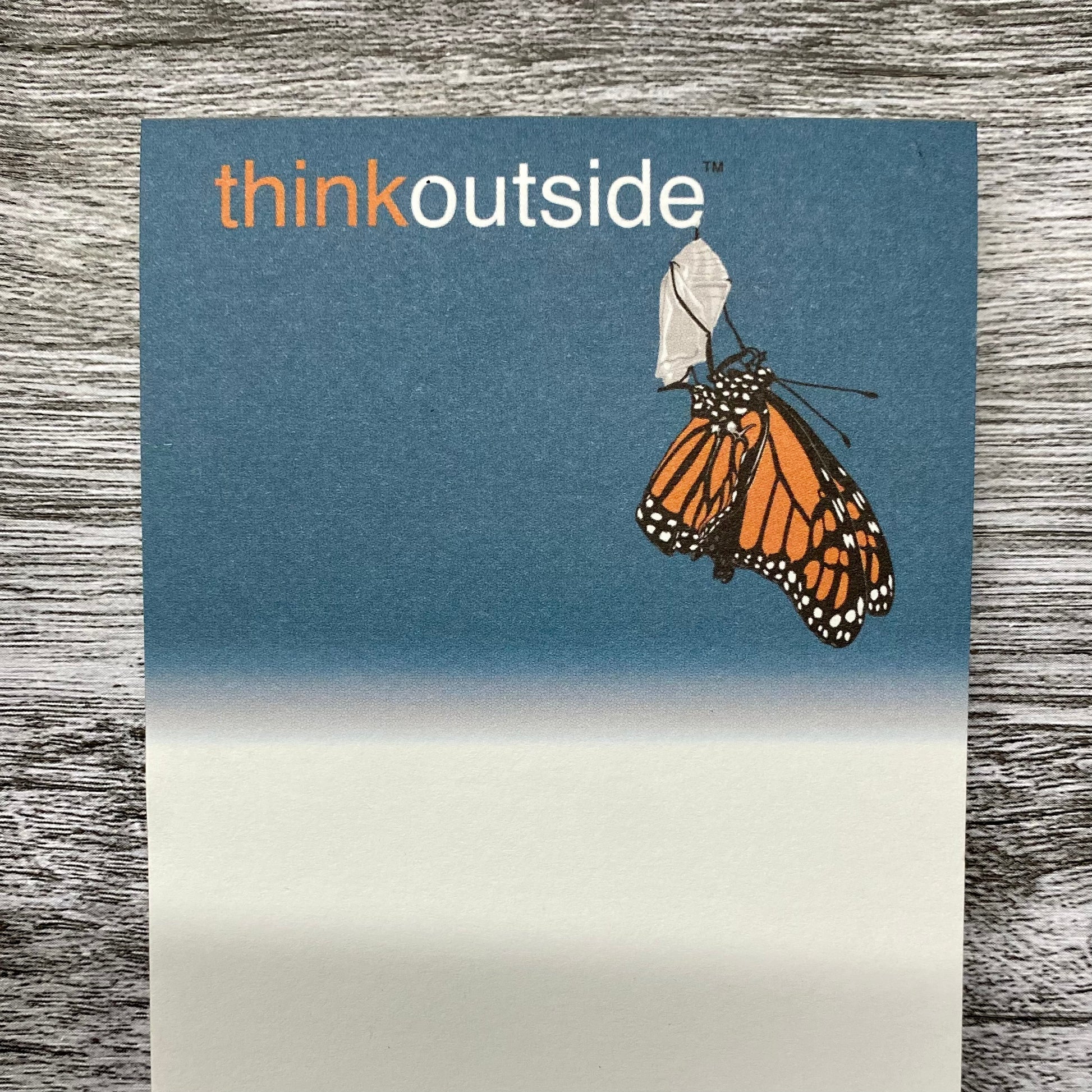 A notepad of 50 sheets of paper featuring a drawing of a monarch butterfly hanging upside down clutching onto its shell.. It also includes the phrase Think Outside